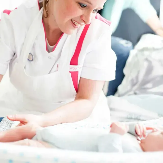 Een verpleegster in een wit uniform glimlacht terwijl ze naar een baby in een wiegje kijkt.