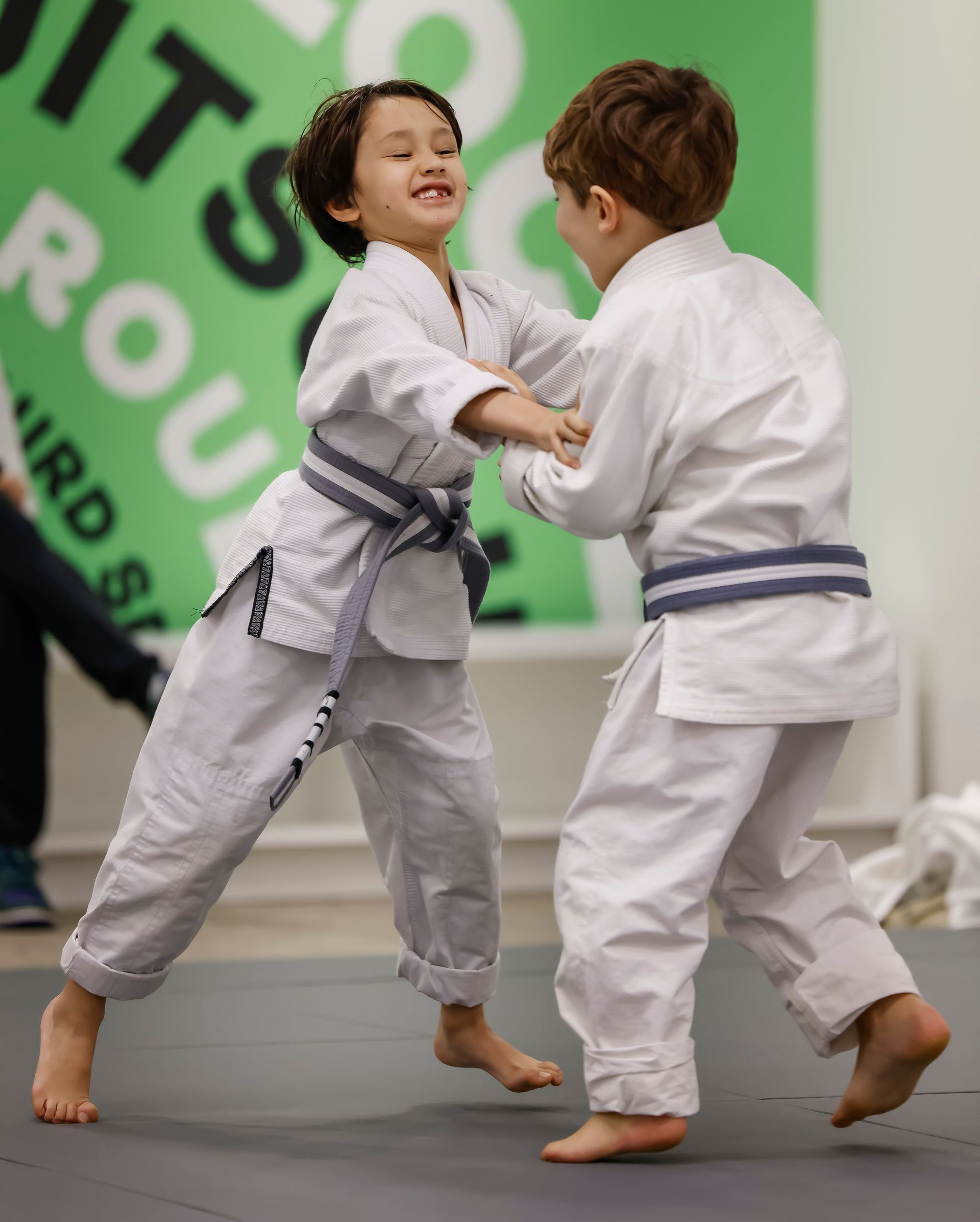 Two children in white martial arts uniforms practice on a mat, smiling and grappling.