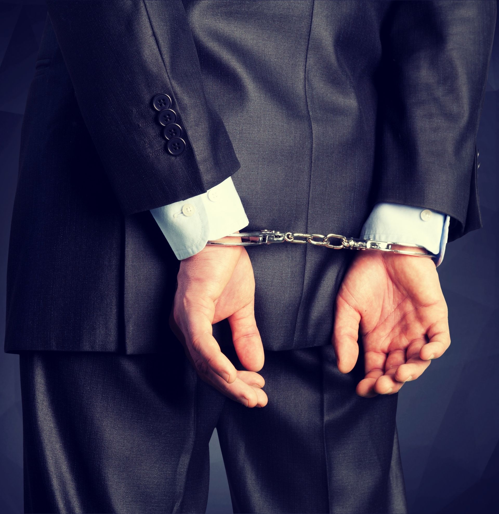 A person wearing a dark suit with their hands restrained behind their back by handcuffs.