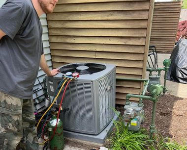 A person in a gray shirt and camouflage pants uses manifold gauges to service an outdoor air conditioning unit.