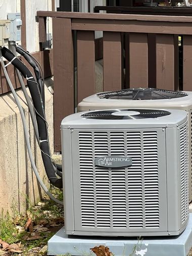 Two silver outdoor central air conditioning units sit on concrete pads beside a house with brown wooden railings.