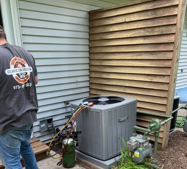 A technician wearing a work shirt connects gauges to an outdoor AC unit next to a wooden privacy fence and gas meter.