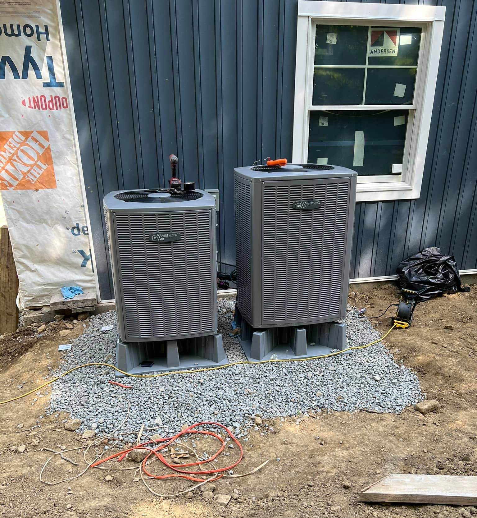 Two grey HVAC units sit on a gravel pad outside a house with blue vertical siding and a window.