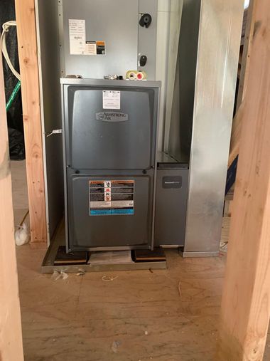 A dark grey HVAC furnace unit sits on a metal base in an unfinished room framed by wooden studs and plywood flooring.