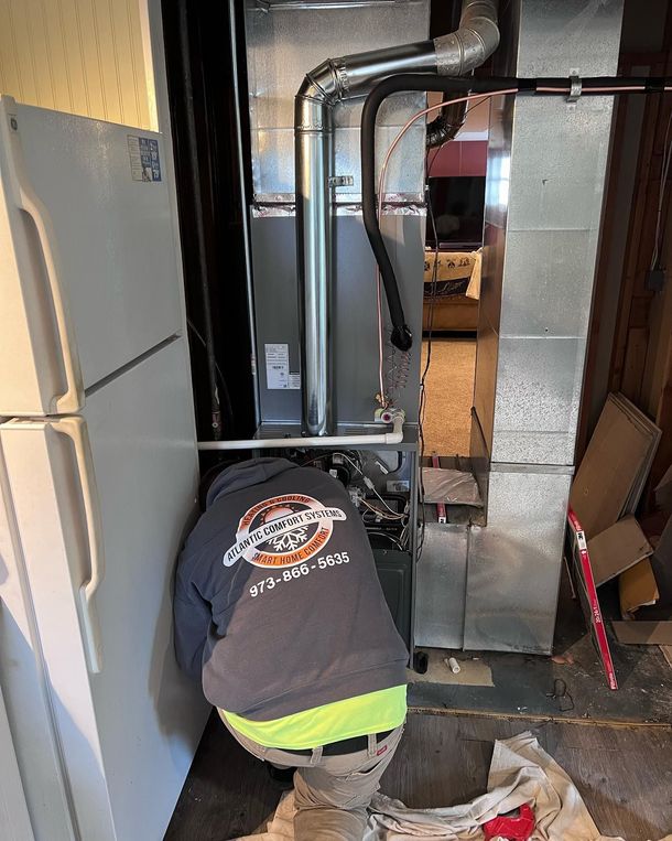 A technician in a branded hoodie kneels while repairing an HVAC unit next to a white refrigerator in a utility room.