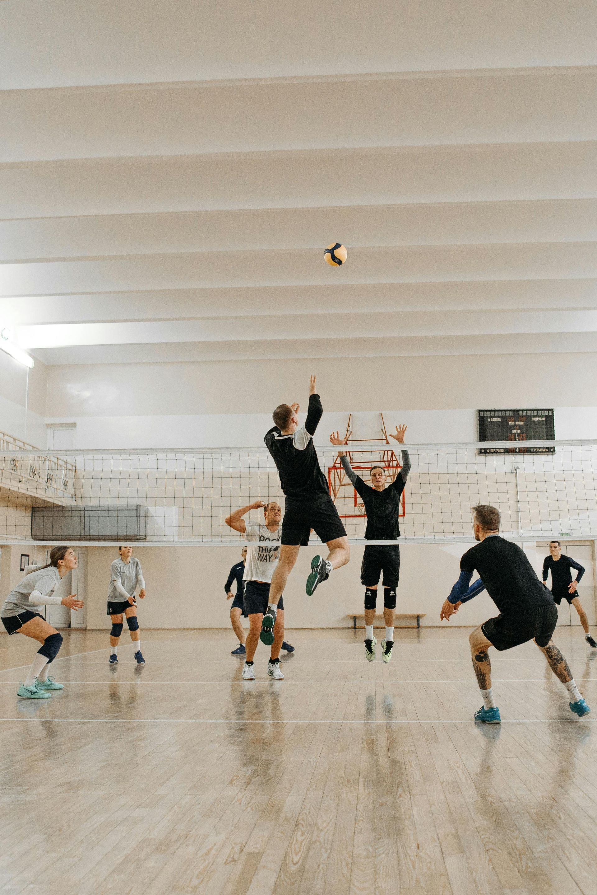 Athletes in an indoor court play volleyball, with one player jumping to strike the ball over the net.