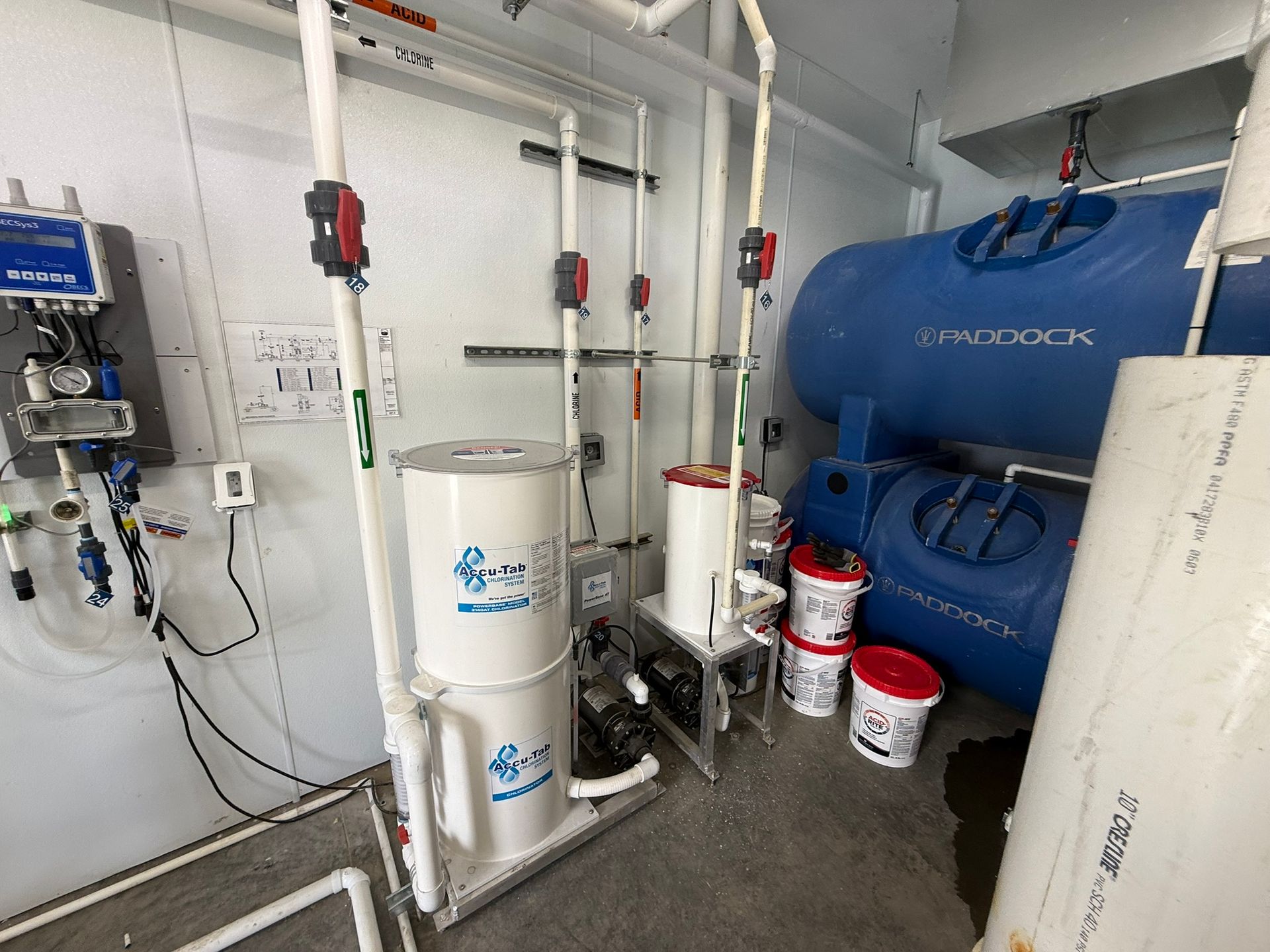 Pool equipment room with white pipes, blue tanks, and chemical containers.