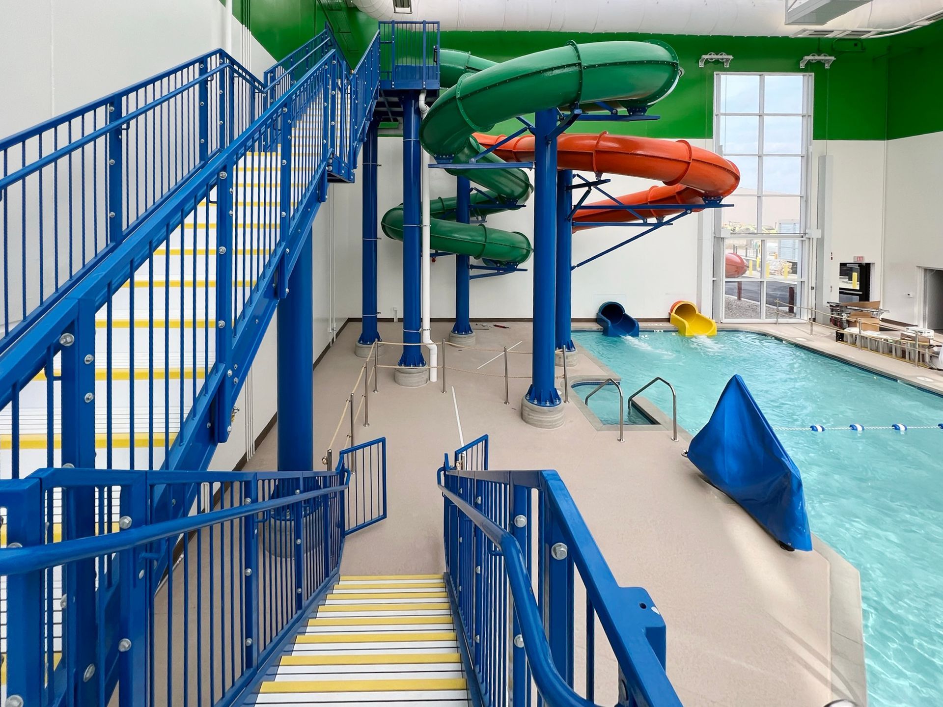 Blue staircase to water slides at an indoor pool. Green, orange, and yellow slides descend into the water.