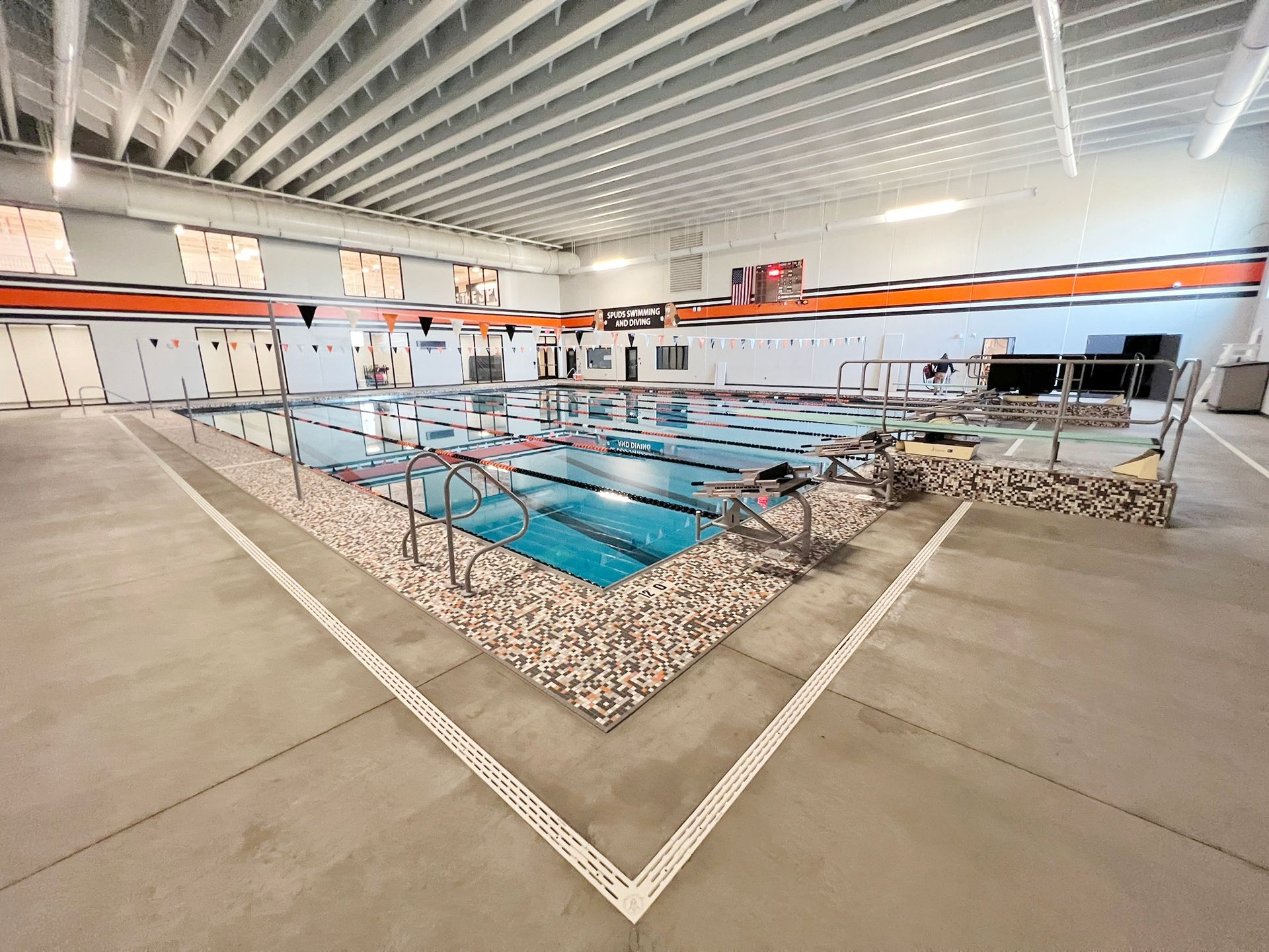 Indoor swimming pool with swimmers, surrounded by a pebble-lined deck, under a white ceiling.