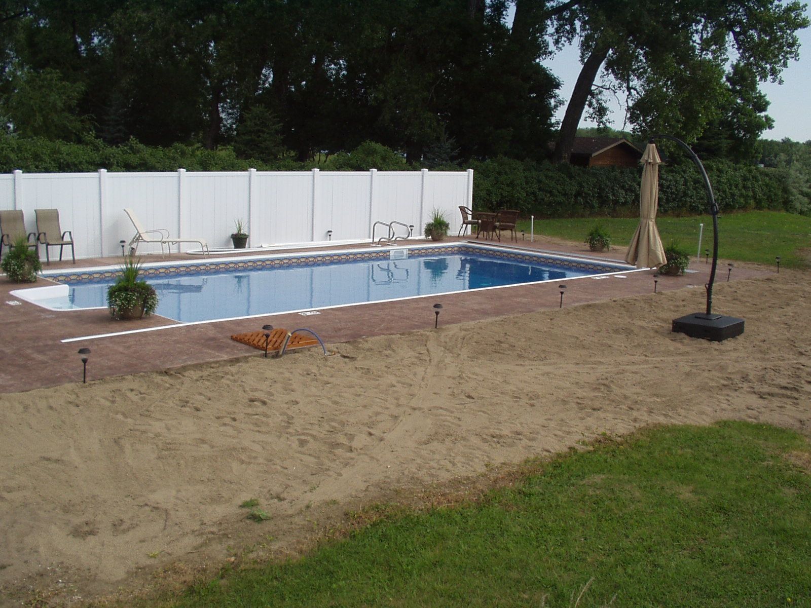 Rectangular pool with white fence, lounge chairs, and umbrella in a backyard setting.