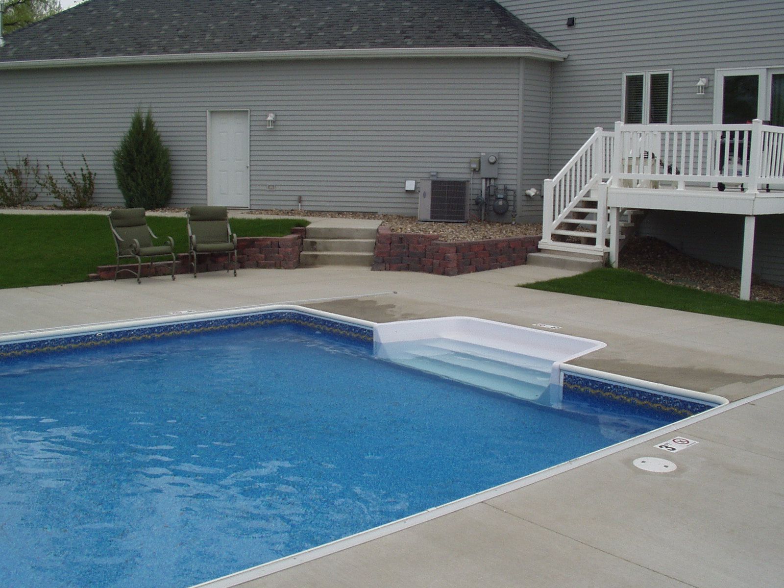 Swimming pool with attached steps and deck. House and landscaping in the background.