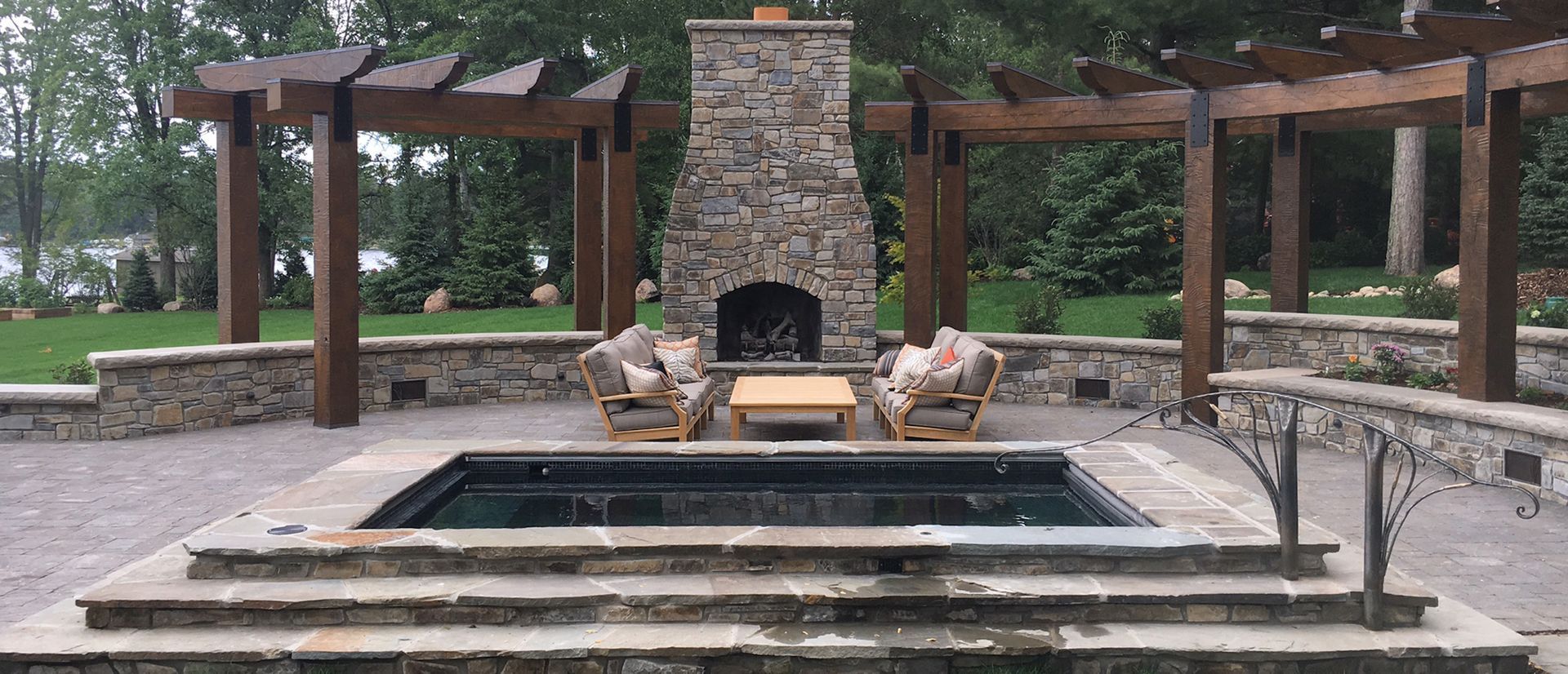 Outdoor living space with a stone fireplace, seating, and a hot tub. Pergola overhead.