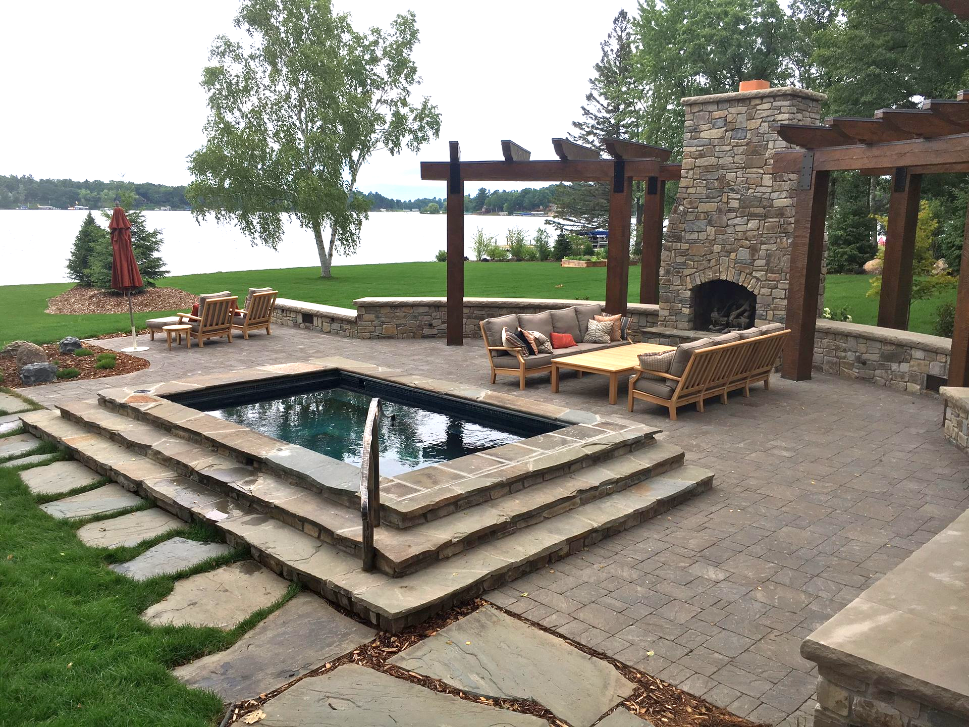 Stone patio with hot tub, fireplace, pergola, and lake view.