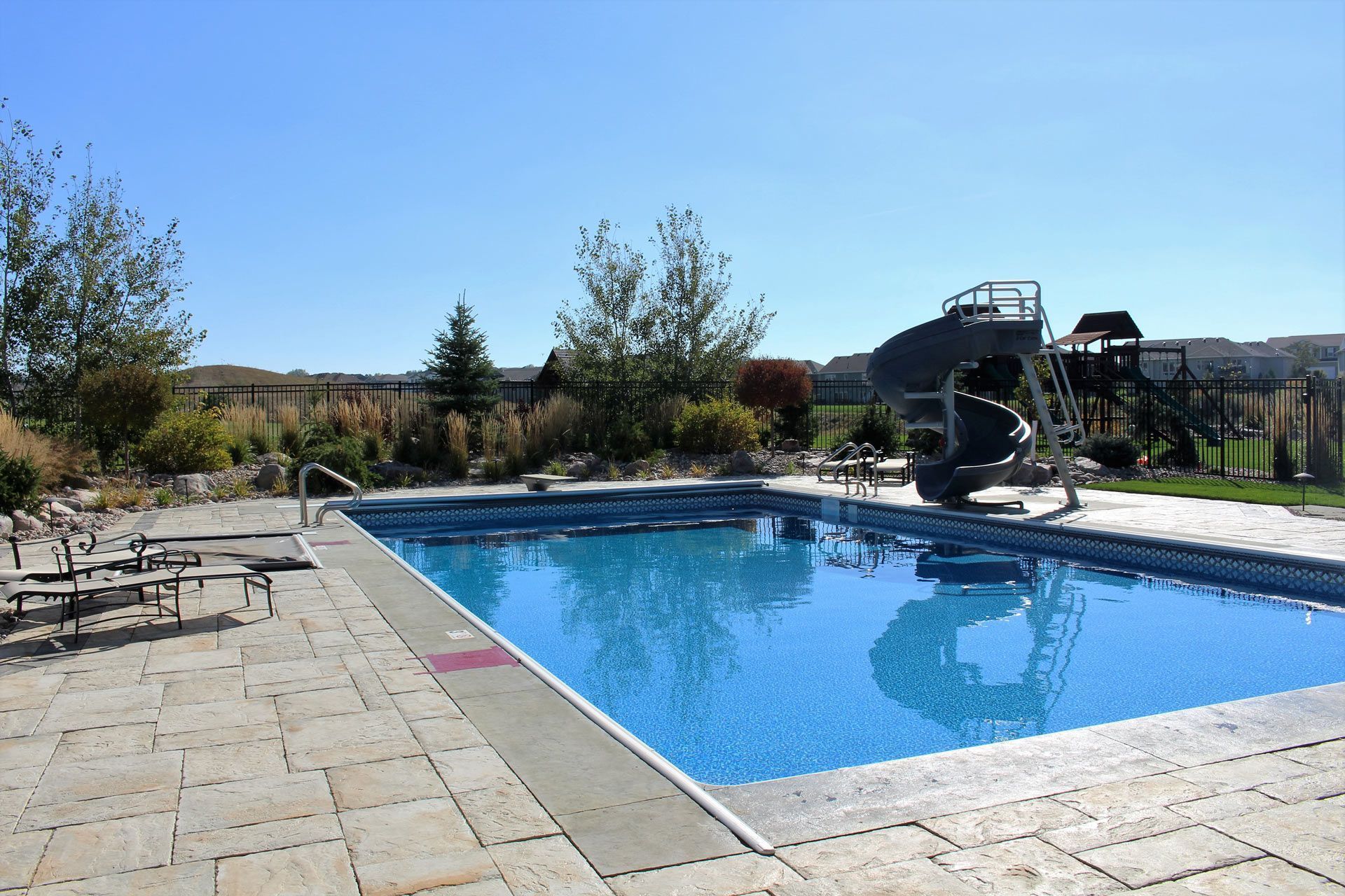 Pool with a slide on a sunny day; chairs on patio, green grass, trees.