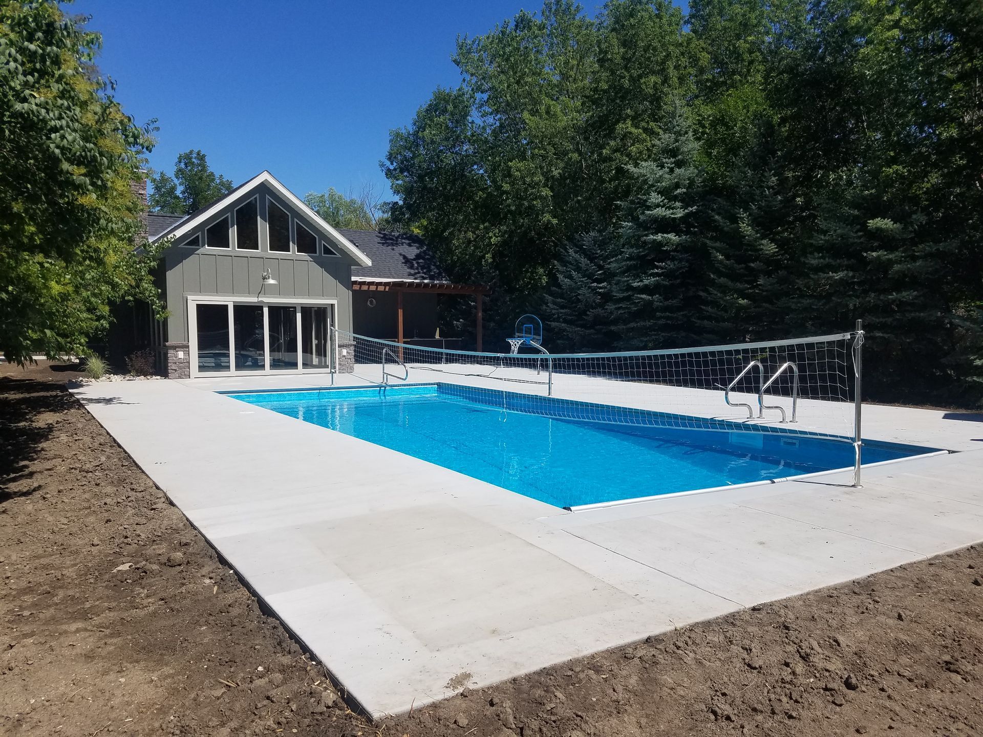Pool with concrete deck, volleyball net, and a house in a grassy setting on a sunny day.