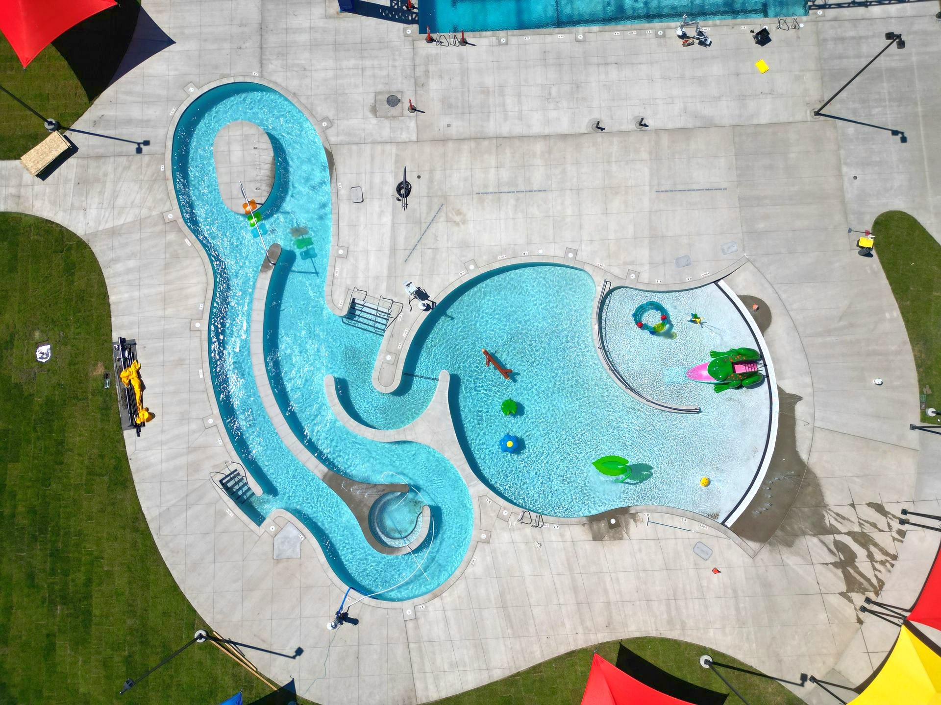 Aerial view of a unique-shaped pool with water features, surrounded by concrete, grass, and colorful shade structures.
