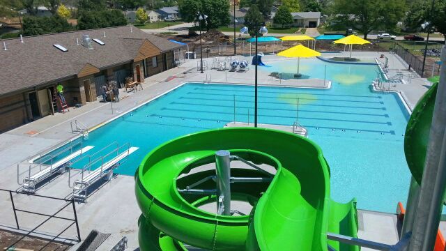 Green water slide overlooking a blue pool with diving boards and a building.