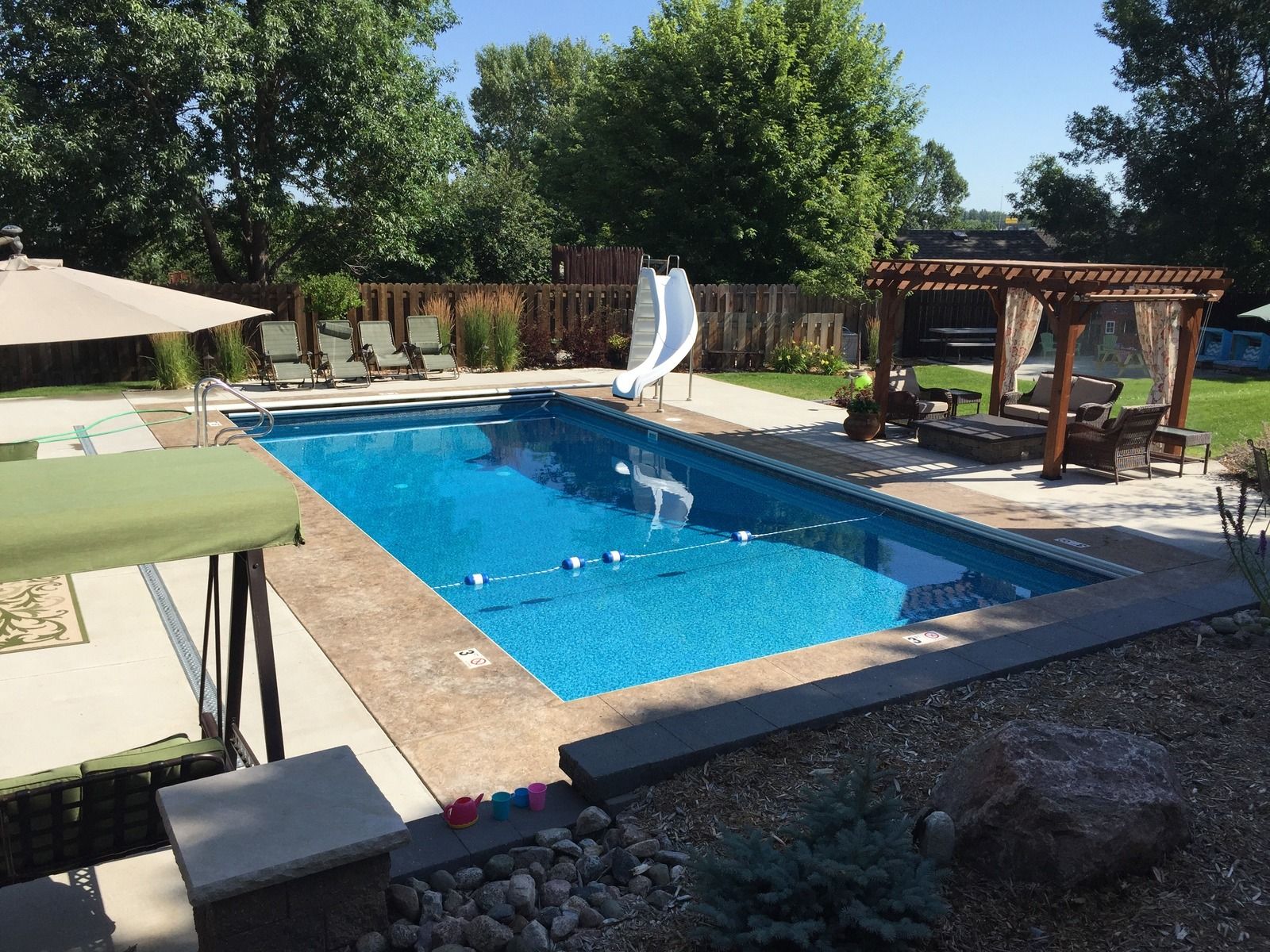 Rectangular swimming pool with slide, patio furniture, and a pergola in a backyard on a sunny day.