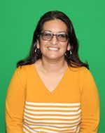 A woman wearing glasses and a yellow sweater is smiling in front of a green background.