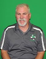 A man with a beard and a gray shirt is sitting in front of a green background.