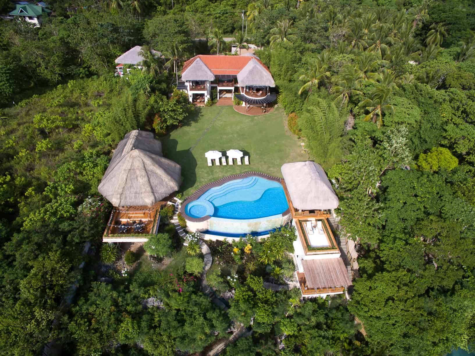 Aerial view of a tropical villa with a pool, grassy lawn, and thatched roof structures surrounded by lush green vegetation.