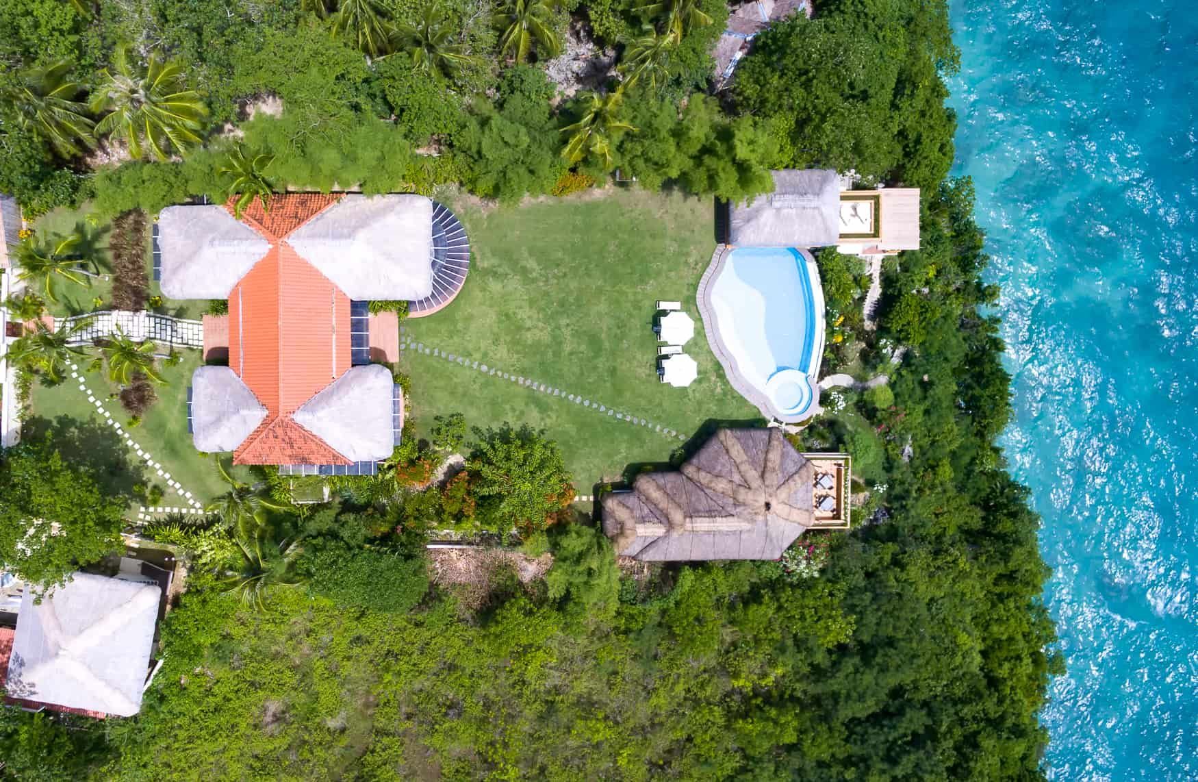 Aerial view: Luxury villa with a pool and oceanfront. Green grass, turquoise water, and lush vegetation surround the buildings.