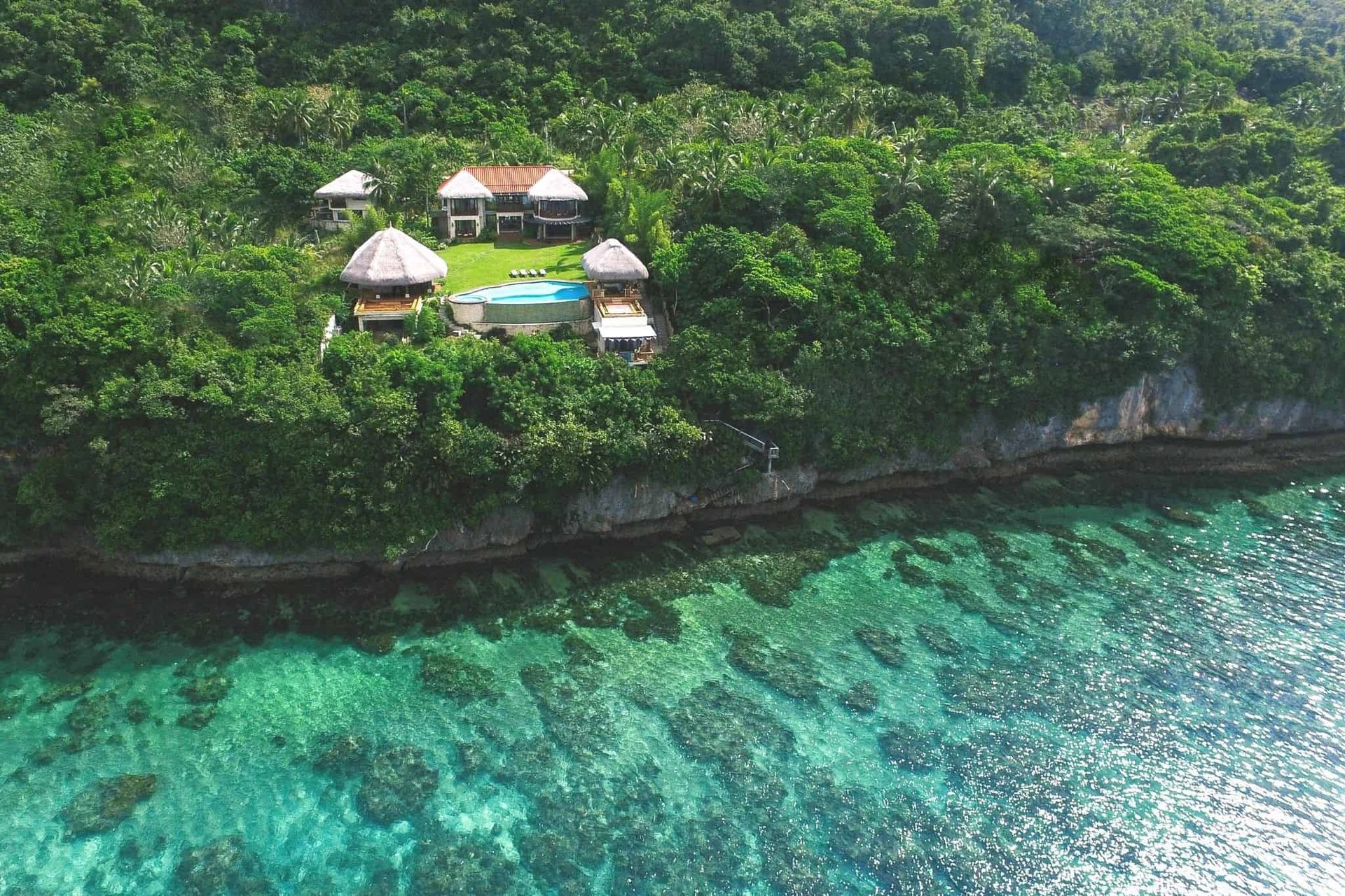 Aerial view of a coastal house with a pool, surrounded by lush green trees and turquoise ocean water.