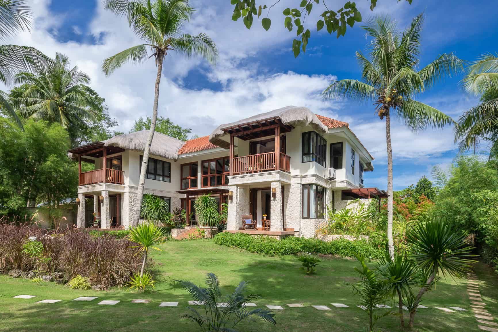 Two-story white house with balconies, red tile roof, and palm trees, surrounded by green grass and tropical plants.