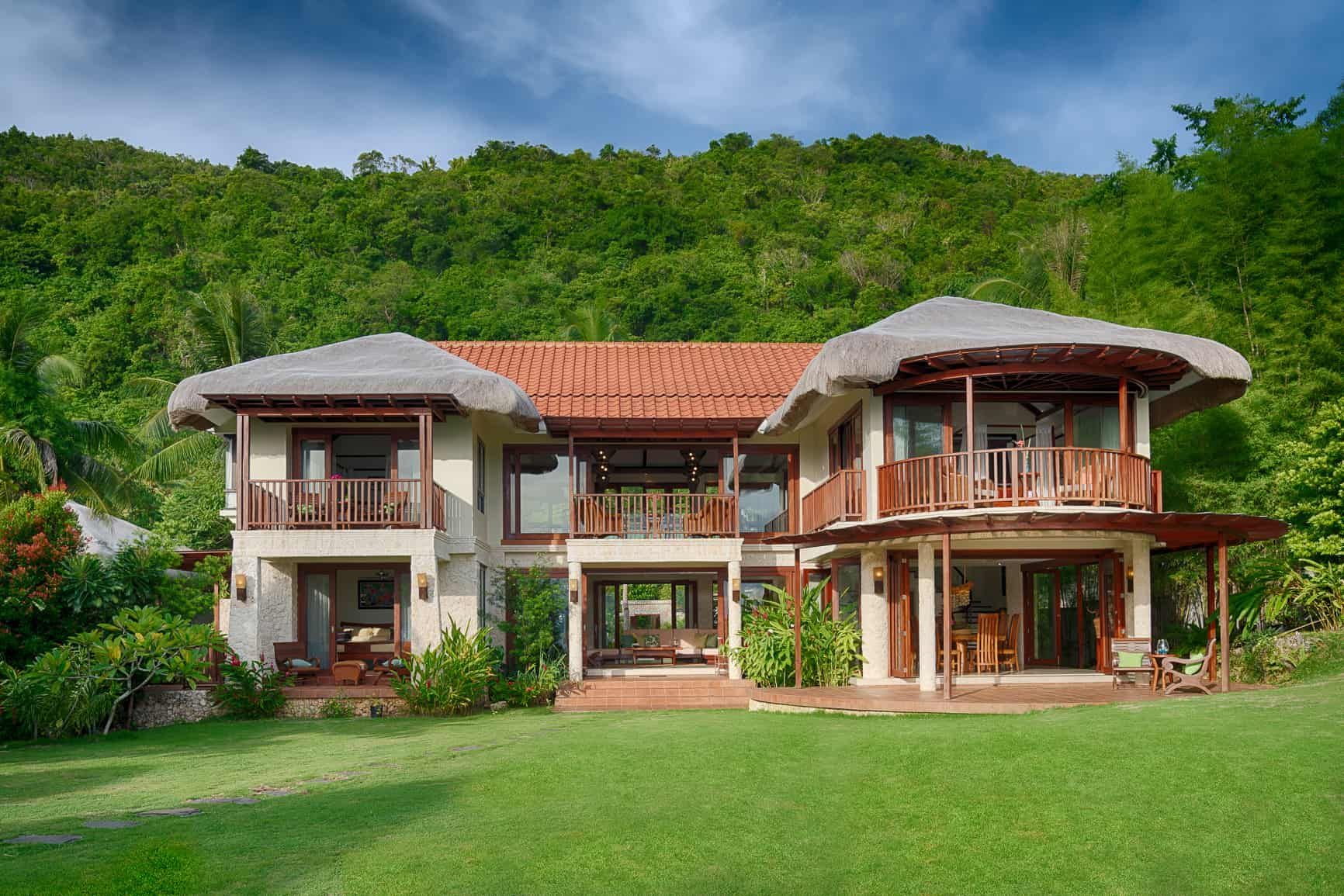 Two-story tropical house with thatched roofs and red tile, surrounded by green lawn and lush vegetation.