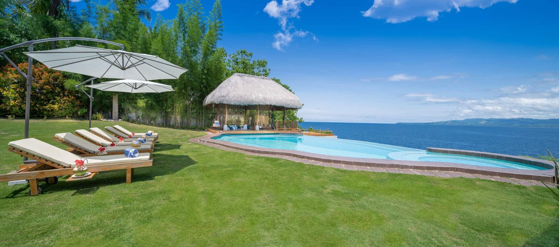 Lush green lawn with pool overlooking ocean. Lounge chairs under umbrellas, thatched roof gazebo in distance. Sunny, blue sky.