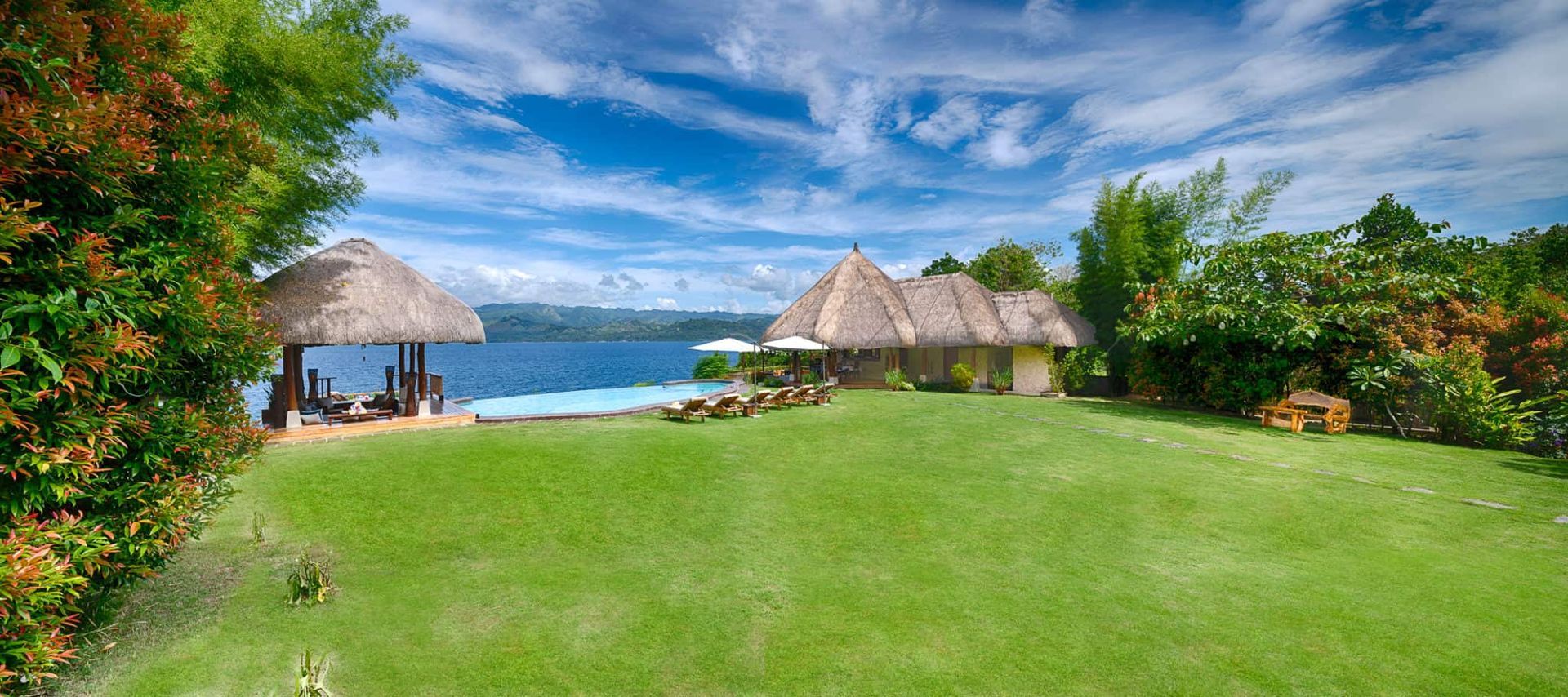 Tropical villa on a lush green lawn, overlooking a blue lake under a partly cloudy sky.