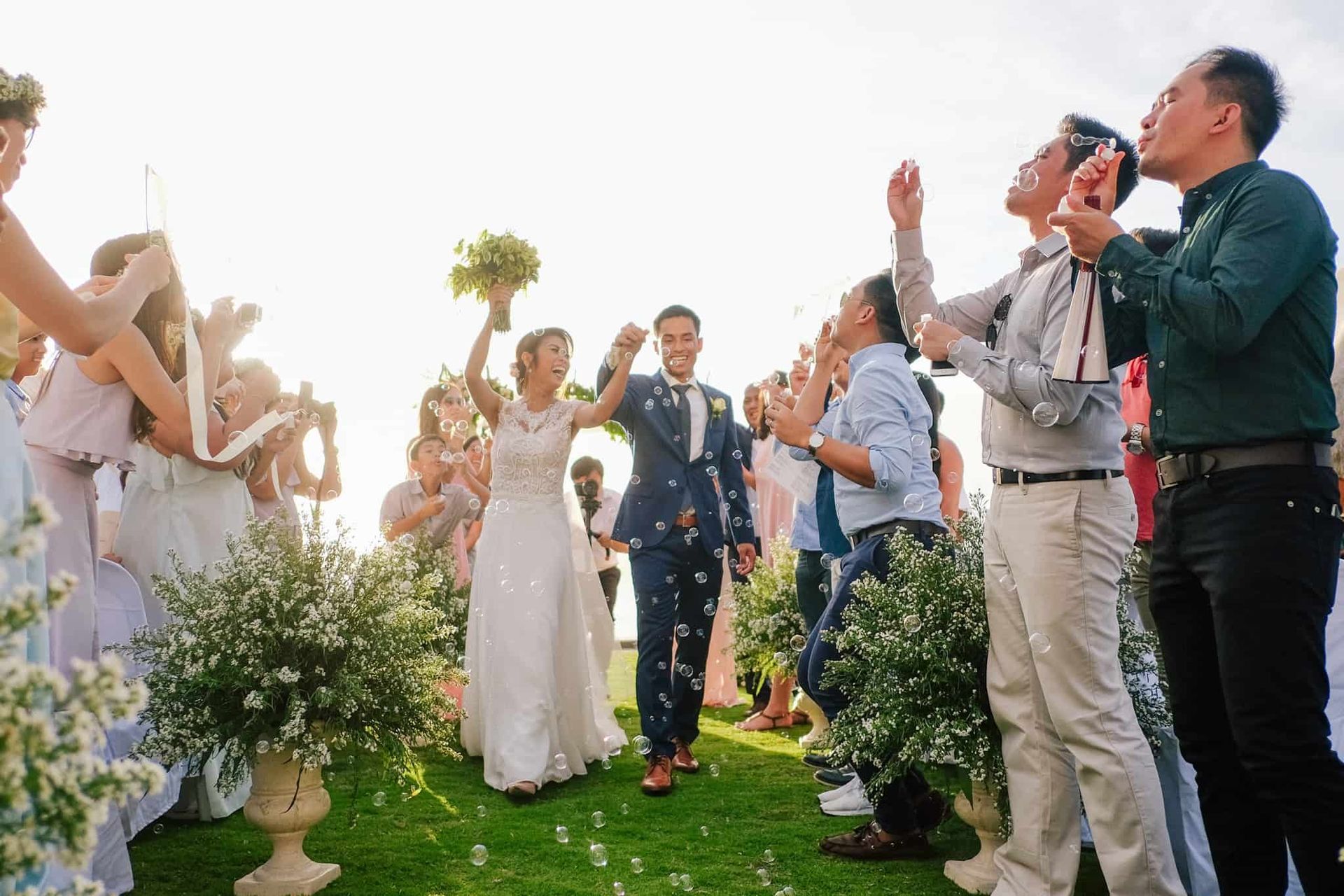 Newlyweds walk down aisle as guests blow bubbles. Sunny outdoor wedding.