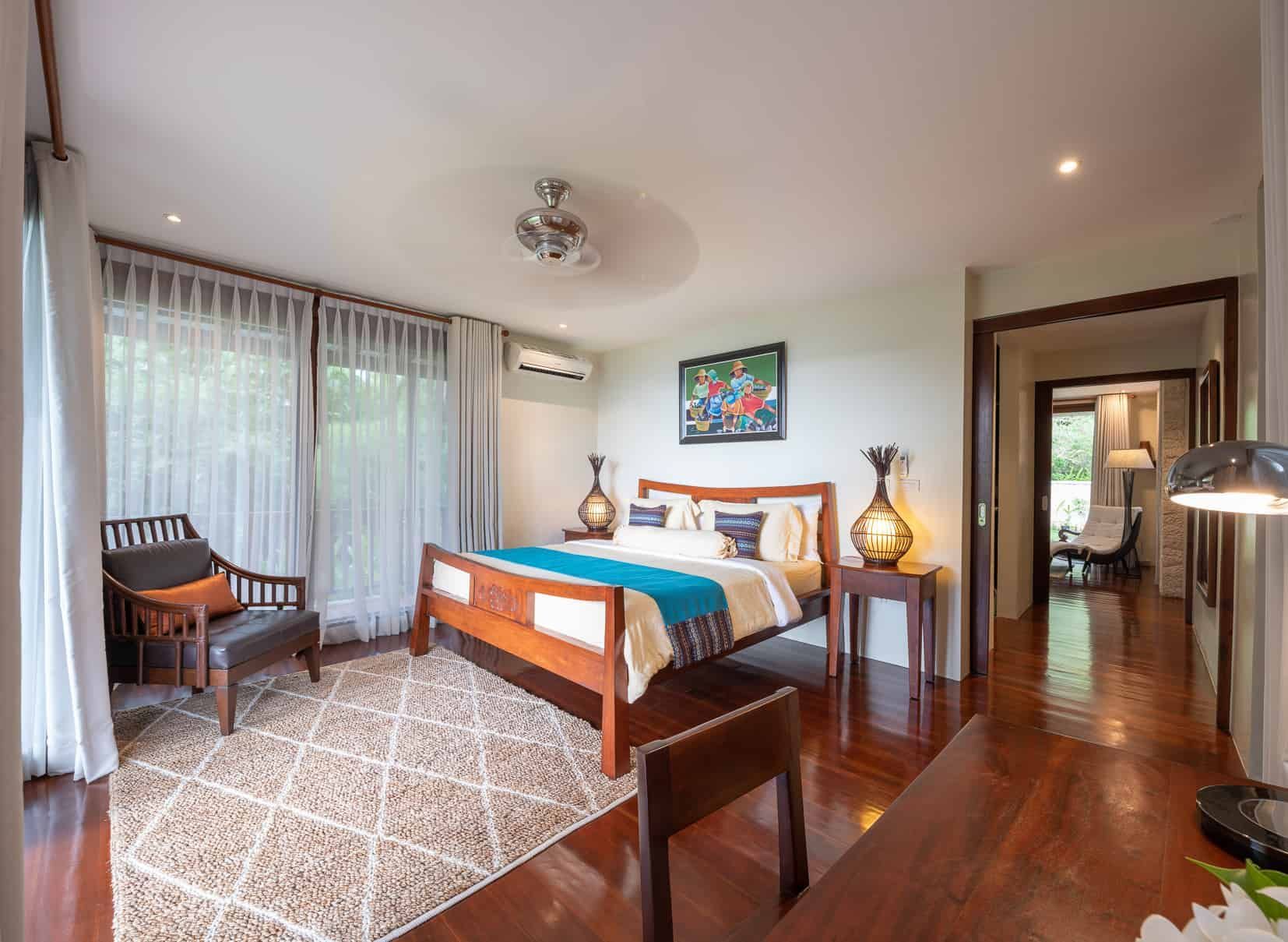 Bedroom with wooden bed, chair, and desk. Large windows and a view to another room.