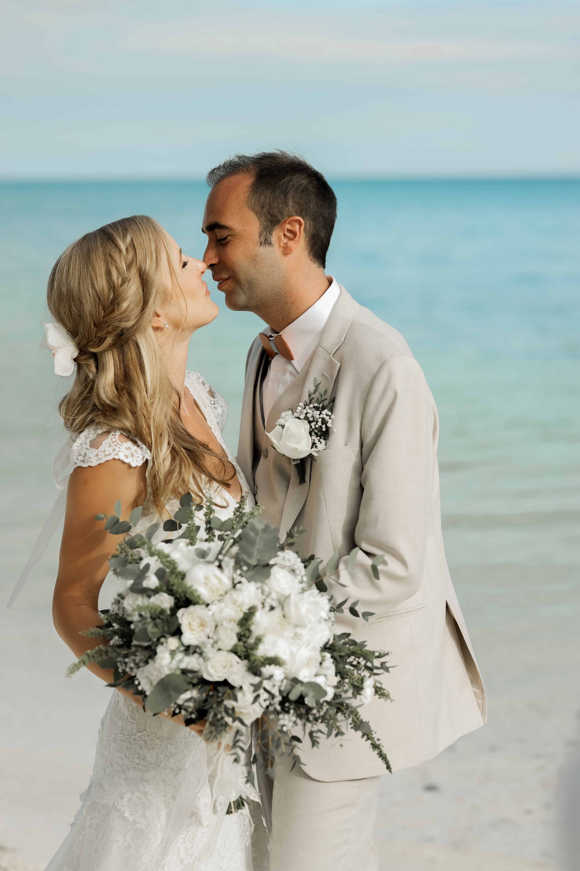 Couple kissing on a beach; the bride holds a bouquet, the groom wears a tan suit. Turquoise water and a light sky in the background.