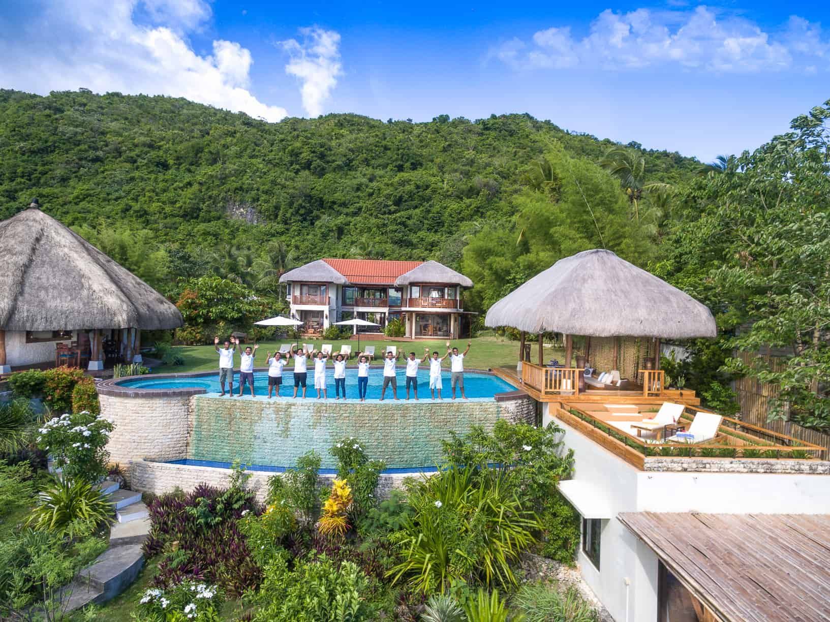 Resort pool with staff in front of multi-story building and thatched roof structures, lush green foliage.