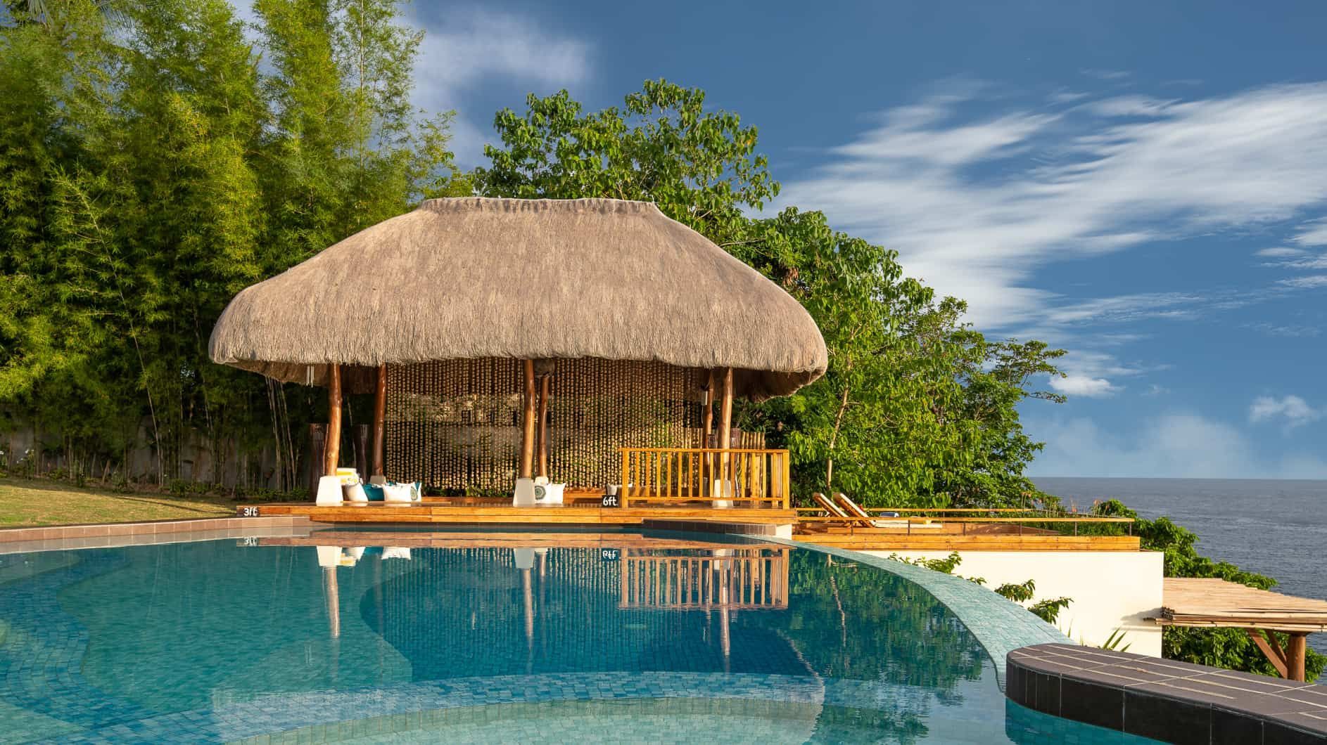 Infinity pool with thatched roof structure overlooking the ocean, set against a bright sky.