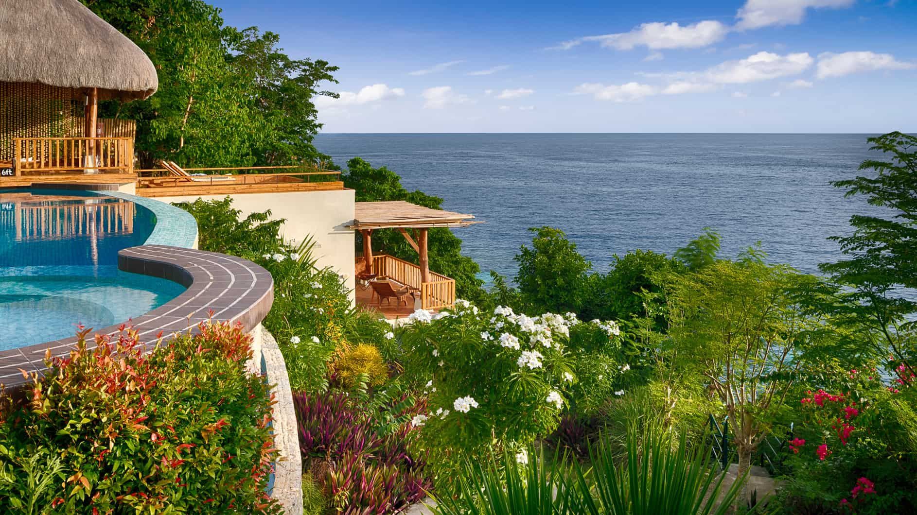 Infinity pool overlooking the ocean, lush gardens, and a thatched-roof structure.
