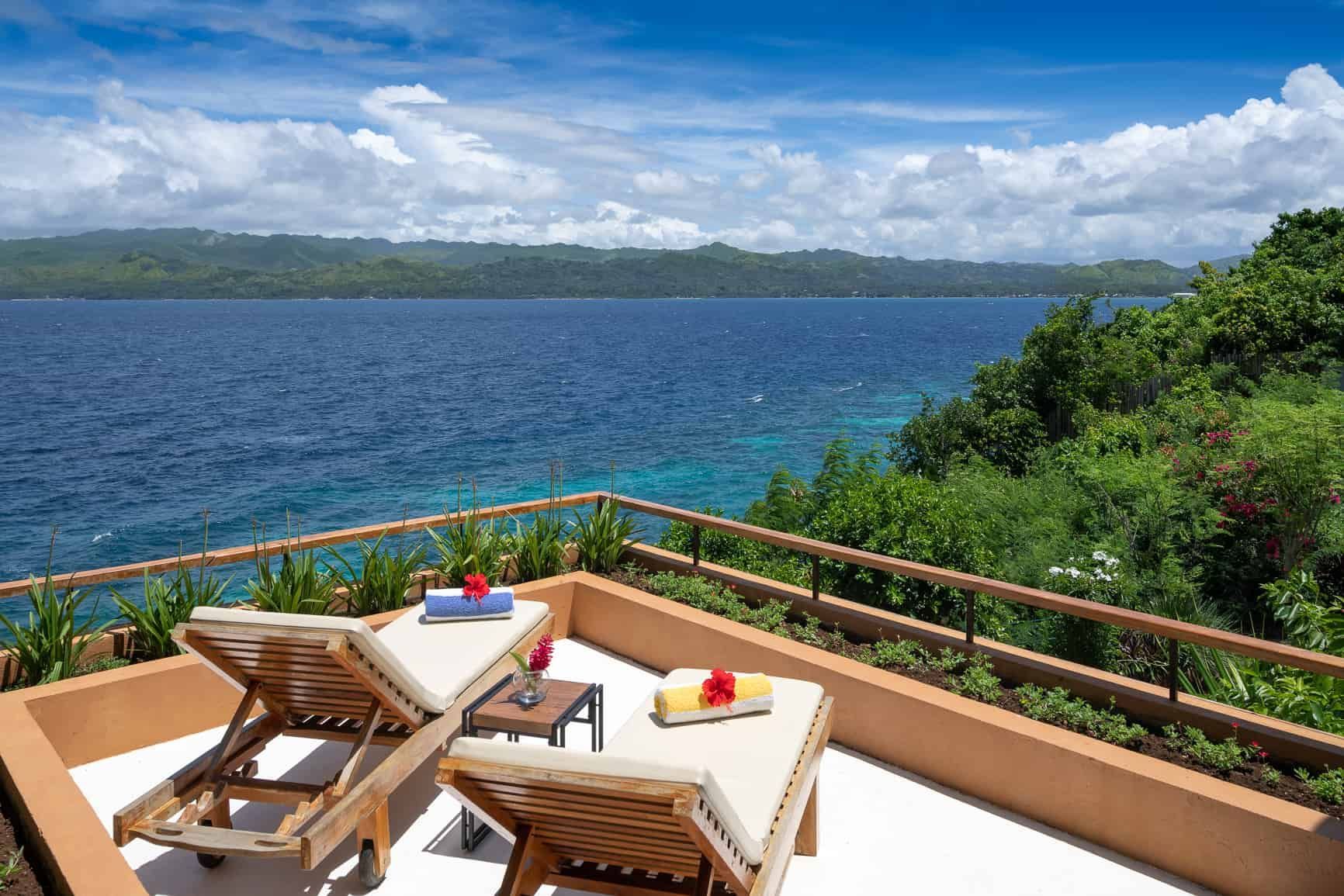 Two lounge chairs on a terrace overlooking a blue ocean, lush greenery, and a cloudy sky.