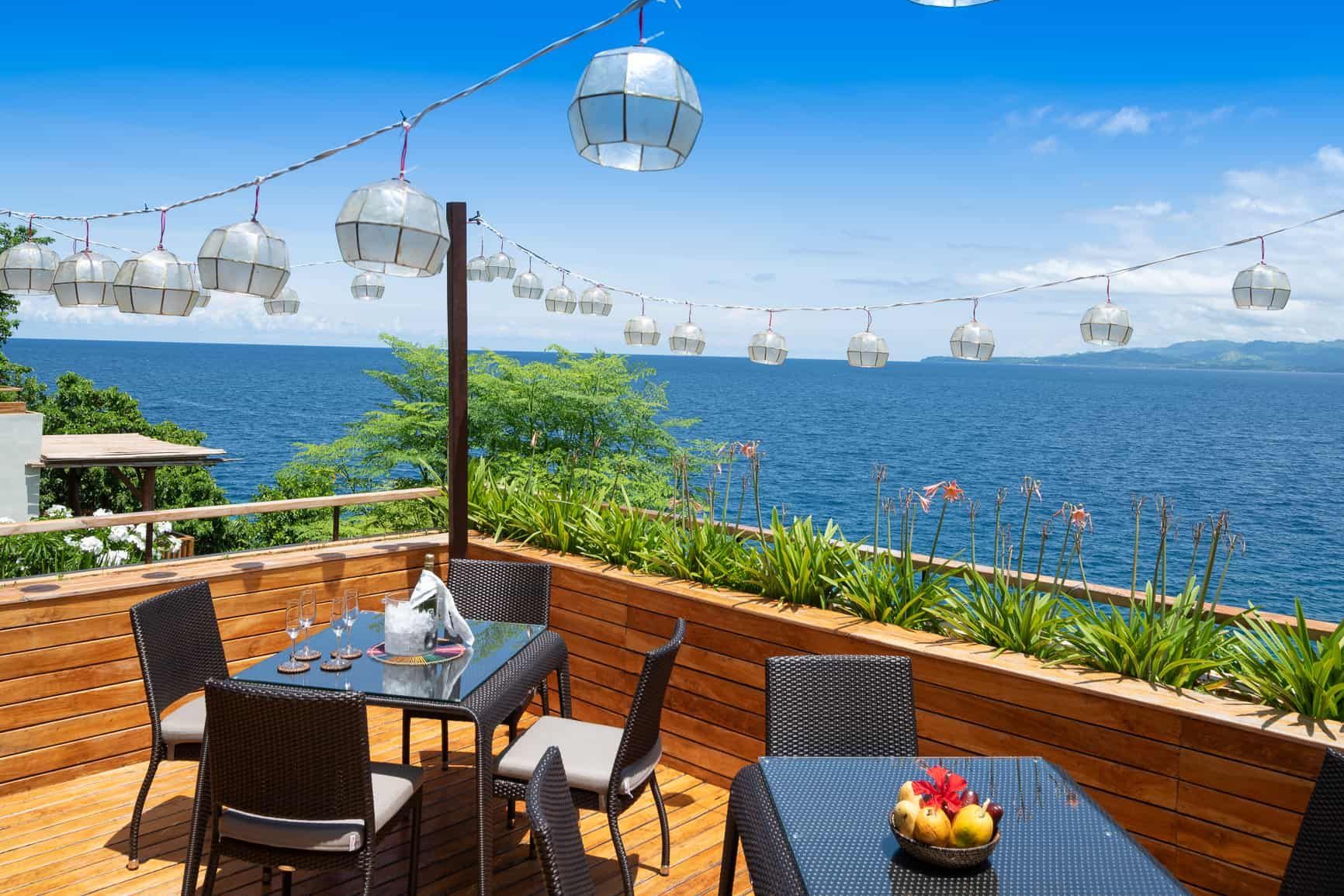 Outdoor restaurant overlooking ocean, tables set with food, string lights.