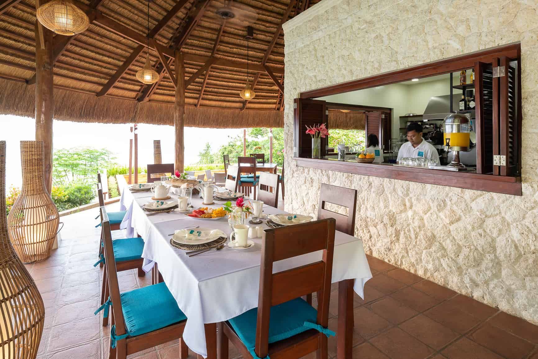 Outdoor dining area with a long table set for a meal, overlooking a view; kitchen window visible.