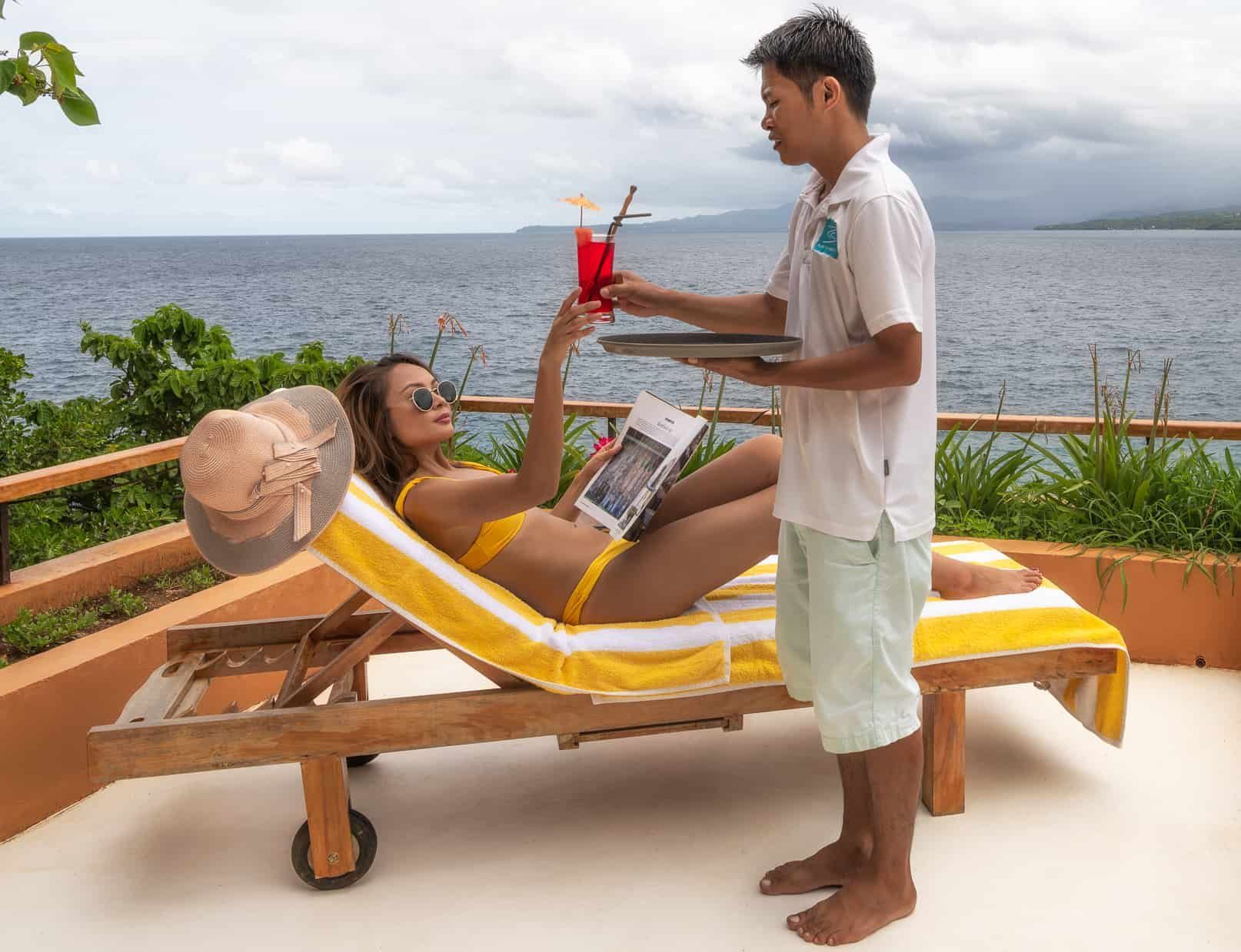 Woman in yellow bikini on lounge chair receives cocktail from server on a deck overlooking ocean.