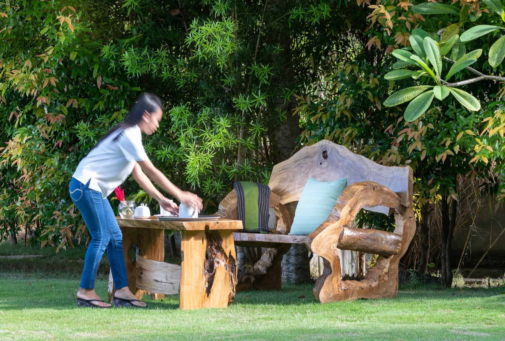 Woman setting a table with a teapot and cups on a wooden table near a rustic wooden bench in a garden.