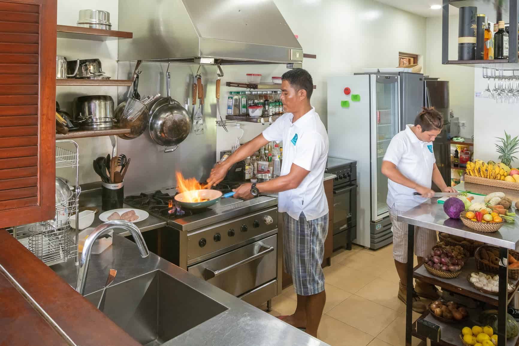 Two people cook in a kitchen with a stove on fire. One stirs a pan, the other cuts vegetables.