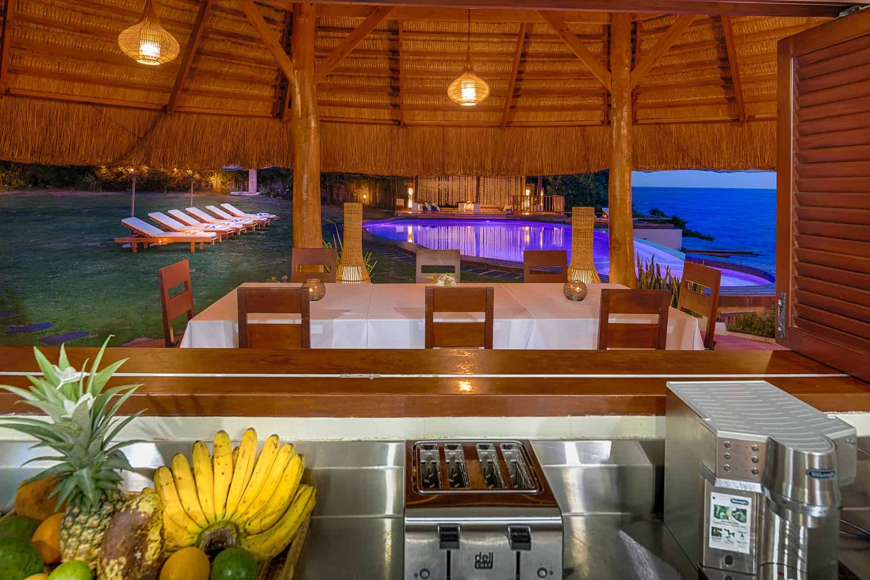 Tropical dining pavilion with a beach view, table set for a meal, a fruit and kitchen counter.