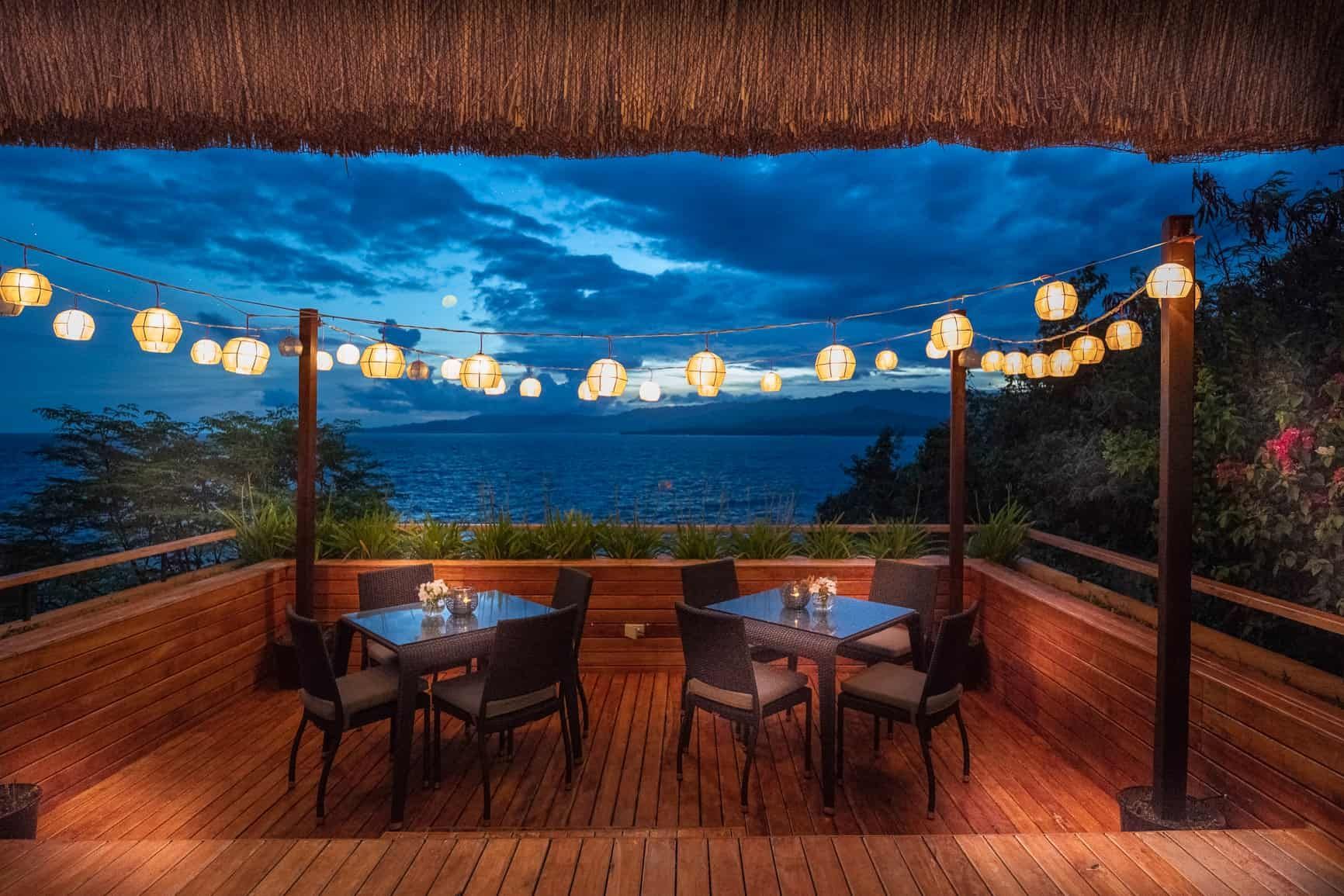 Wooden deck restaurant overlooking a calm ocean at dusk. Lanterns and tables with chairs set.