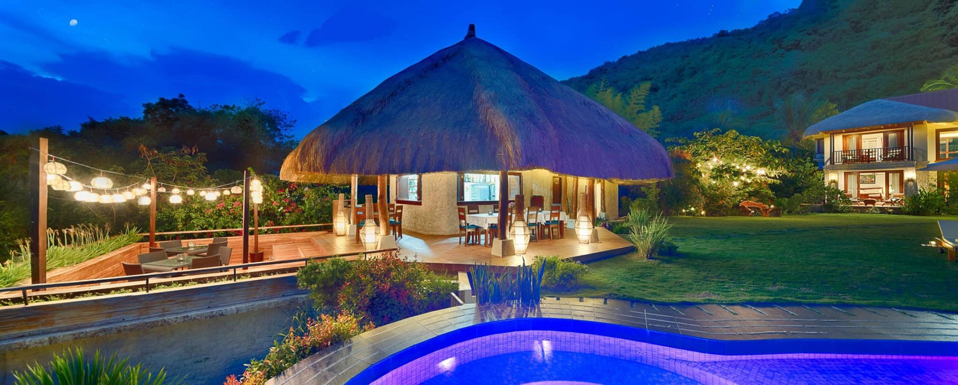 A resort scene at dusk featuring a thatched-roof structure, a pool, and illuminated buildings.