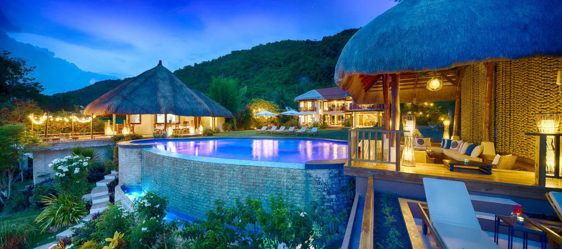 Lush tropical resort with a pool overlooking a mountain. Lit by warm lights, cool blue water and sky.