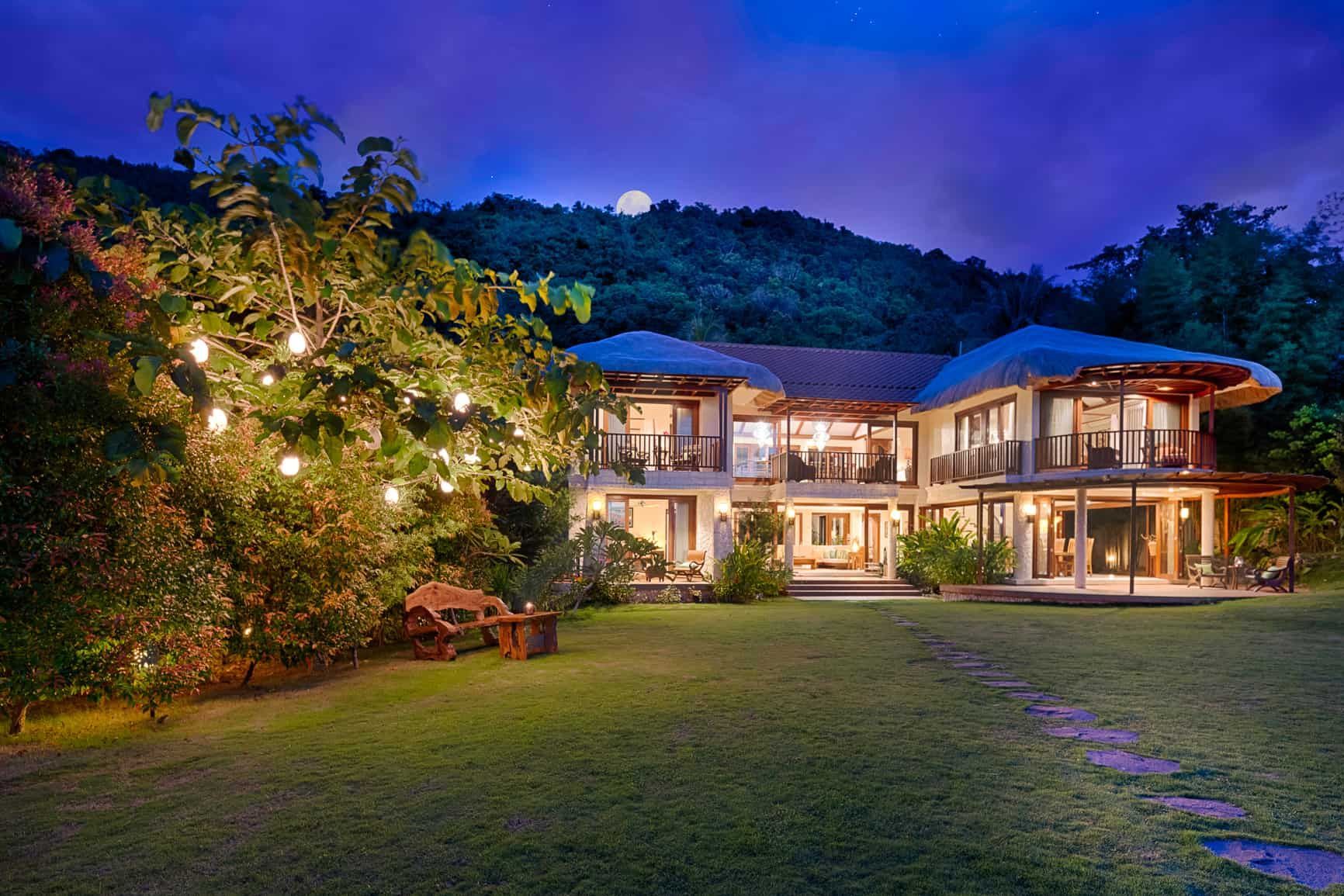 Two-story house with lights, lit up at night, with a mountain backdrop and a full moon.