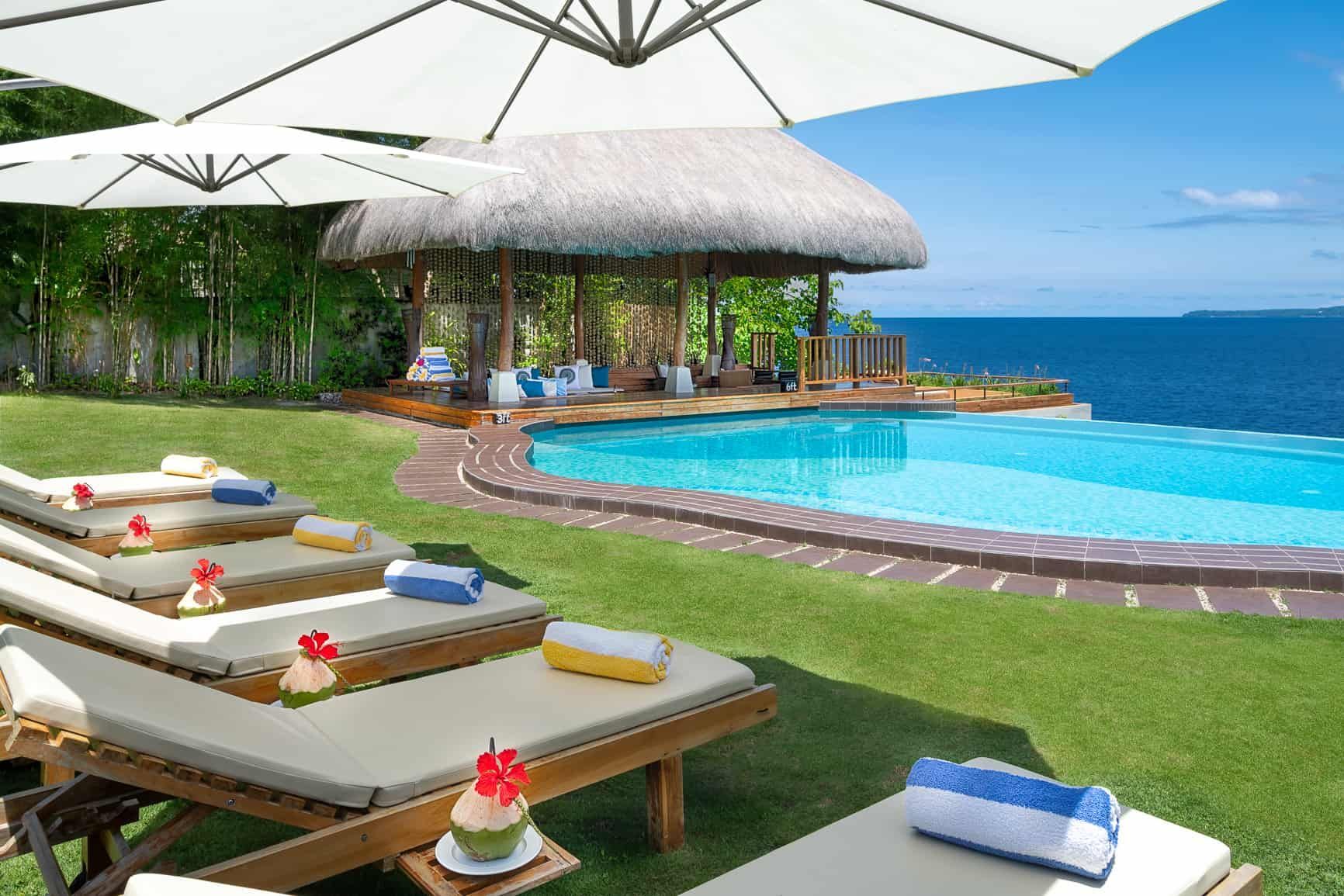 Poolside scene: lounge chairs, umbrellas, infinity pool overlooking ocean, thatched roof hut, sunny day.