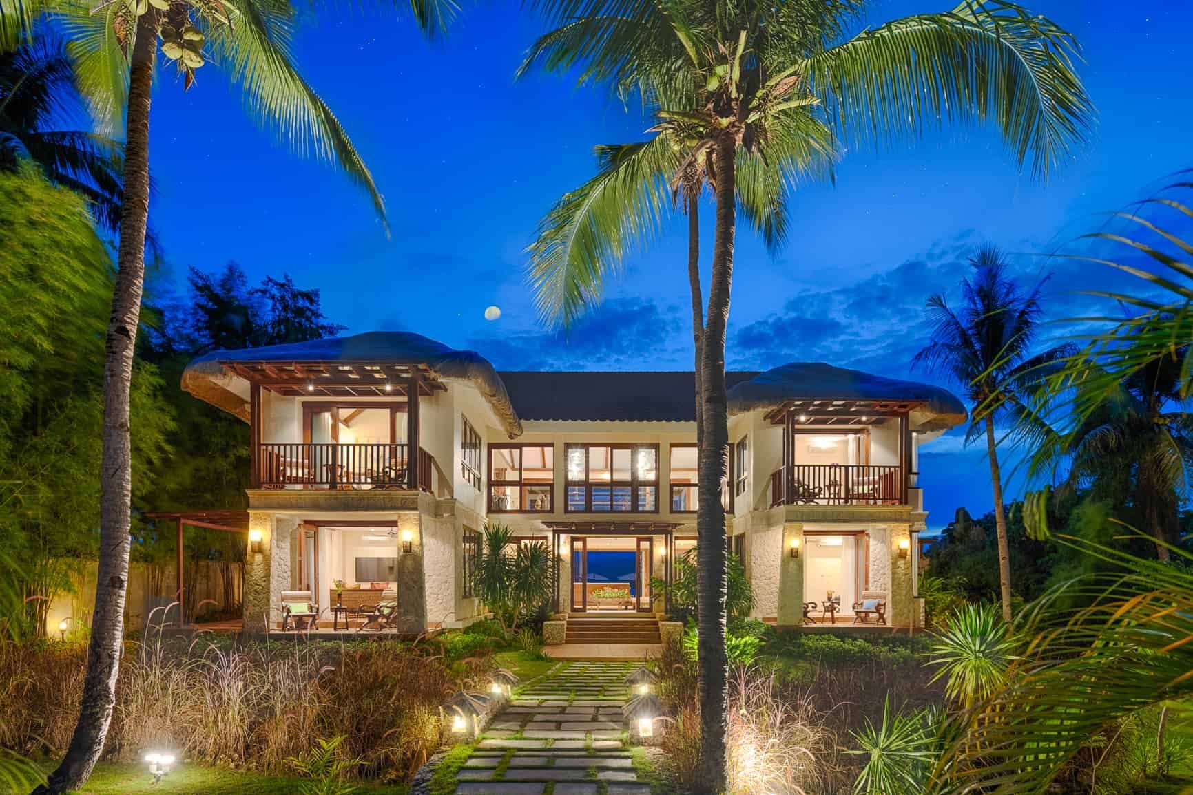 Two-story beachfront villa at night, illuminated, with palm trees and a pathway leading to the entrance.